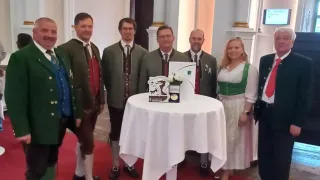 Eine Gruppe von Männern und einer Frau in traditioneller Kleidung steht vor einem Tisch in einem großen Saal. Der Tisch hat eine kleine Schachtel und eine Blume. Sie lächeln.