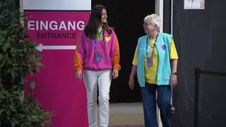 Zwei Frauen gehen Seite an Seite, eine trägt eine rosa Jacke mit einem Herz und die andere ein gelbes Shirt und eine blaue Weste, beide mit Ausweisbändern. Sie stehen vor einem pinkfarbenen Schild mit einem weißen Pfeil und dem Wort 'Eingang'.