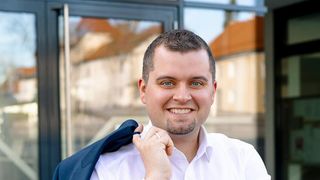 Ein Mann in einem weißen Hemd und einer blauen Anzugsjacke steht draußen und lächelt. Er hält seinen Mantel mit einer Hand am Revers fest.