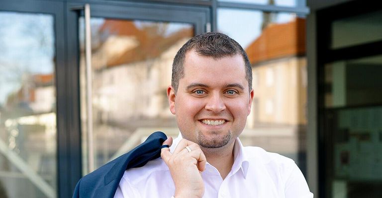 Ein Mann in einem weißen Hemd und einer blauen Anzugsjacke steht draußen und lächelt. Er hält seinen Mantel mit einer Hand am Revers fest.