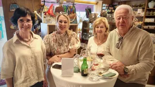 Vier Personen, drei Frauen und ein Mann, lächeln in einem Geschäft. Es gibt einen Tisch mit Wein, einer Karaffe und einem Teller mit Gabel.