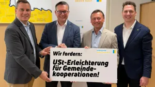 Vier Männer halten ein Schild, das für Steuererleichterungen für Gemeindekooperationen wirbt. Sie sind in Anzügen gekleidet und stehen vor einem Hintergrund mit einer Karte und Logos.