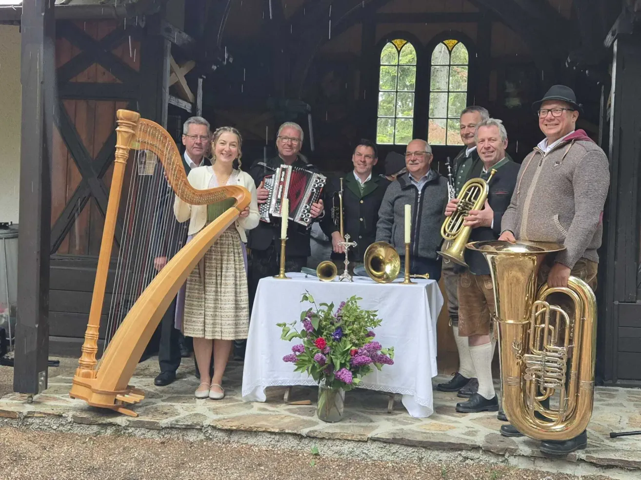 Eine Gruppe von Musikern mit Harfe, Trompeten und Tuba posiert für ein Foto in einem Bauernhaus. Eine Frau hält eine Harfe und lächelt, und ein Tisch mit Blumen und Kerzen steht hinter ihnen.