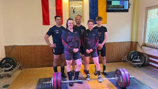 Fünf Personen posieren hinter einer Hantel in einem Fitnessstudio, mit einem Monitor und Fahnen im Hintergrund. Sie tragen Sportbekleidung.