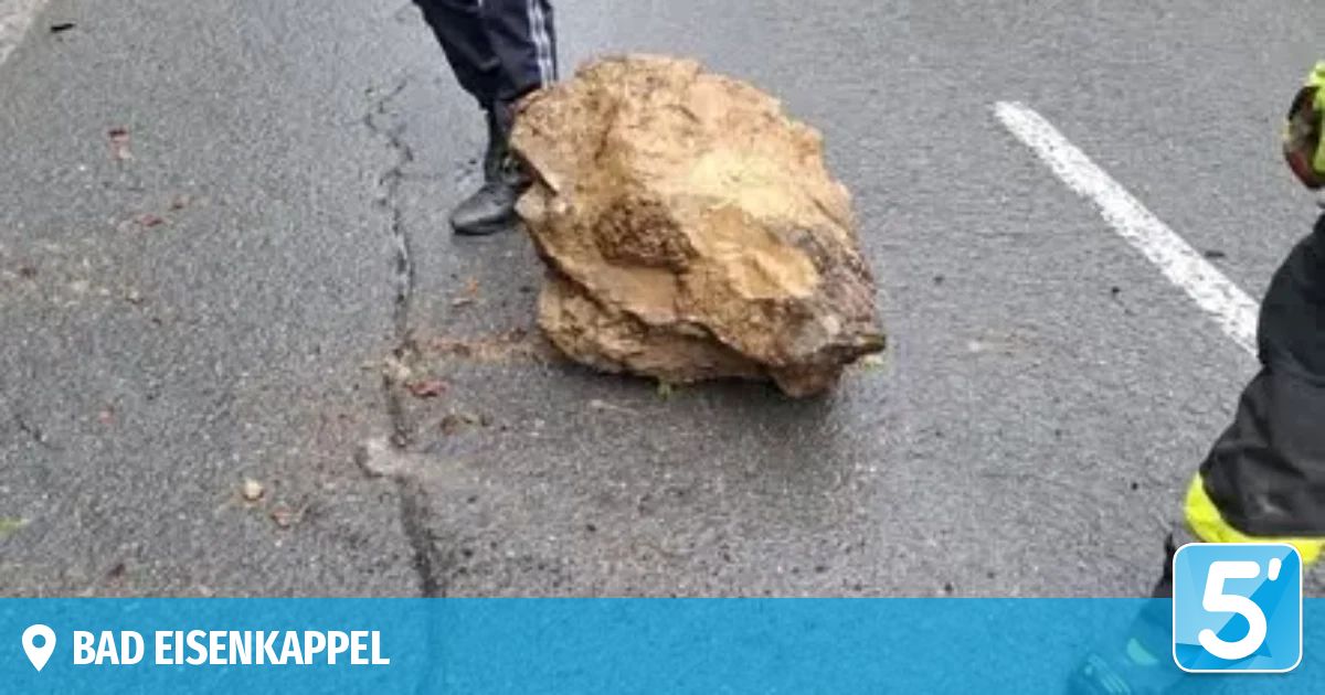 Eine Person steht neben einem großen Stein auf der Straße, teilweise mit Erde bedeckt. Der Stein ist groß und scheint die Straße zu behindern.
