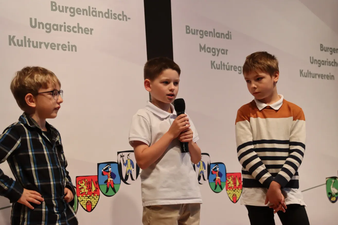 Drei Jungen stehen vor einem Hintergrund mit deutschem Text und Wappensymbolen. Der Junge in der Mitte hält ein Mikrofon.