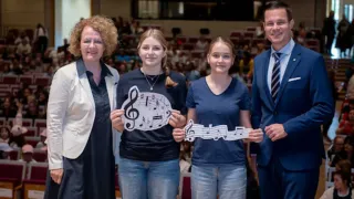 Drei Frauen und ein Mann stehen in einem Raum und halten Musiknoten. Hinter ihnen sitzt ein Publikum auf Bänken. Die Frauen tragen Freizeitkleidung und der Mann einen Anzug mit Krawatte.