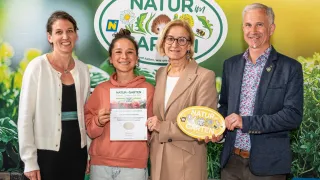 Vier Personen stehen nebeneinander und lächeln für ein Foto. Sie halten ein Zertifikat und einen gelben Abzeichen mit der Aufschrift 'Natur-Garten'. Die Frau auf der linken Seite trägt eine Halskette. Die Frau in der Mitte hält ein Zertifikat. Die Frau auf der rechten Seite trägt eine Brille und einen Ring.