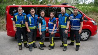 Sechs Feuerwehrleute stehen neben einem roten Einsatzfahrzeug mit Pflanzen und Bäumen im Hintergrund. Sie lächeln alle und tragen blaue Westen.