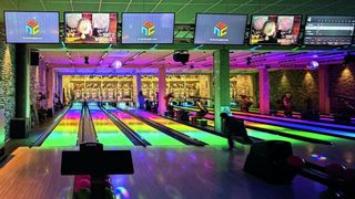 Eine Bowlingbahn mit leuchtenden Lichtern. Monitore hängen von der Decke und zeigen ein Logo. Leute bowlen und genießen die bunte Atmosphäre.