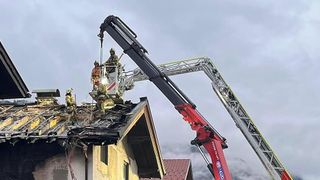 Eine Gruppe von Feuerwehrleuten, die einen Kran benutzen, um Menschen aus einem brennenden Haus zu retten, mit Rauchschwaden und einem roten Dach.
