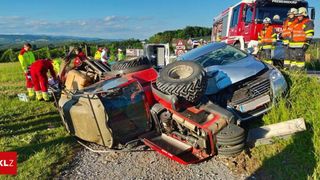 Ein Autounfall mit zwei Fahrzeugen auf einem Schotterweg. Ein Fahrzeug ist umgekippt, und ein anderes ist beschädigt. Feuerwehrwagen sind anwesend, und Leute haben sich versammelt.