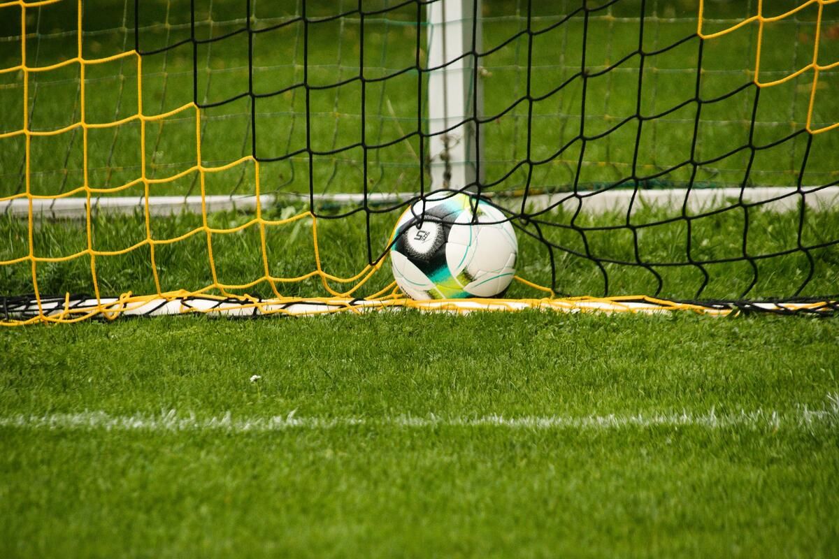 Ein Fußball steht hinter einem Netz auf einem grünrasigen Fußballfeld. Das Netz ist gelb und schwarz.