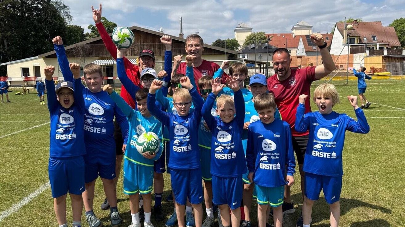 Eine Gruppe Jungen in blauen Trikots posiert mit zwei Fußballen und zwei Männern, einer trägt ein rotes Shirt, unter einem bewölkten Himmel.