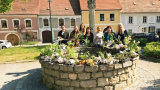 Eine Gruppe von Frauen steht um einen runden Steinpflanzer mit verschiedenen Topfpflanzen. Sie lächeln und scheinen für ein Foto zu posieren. Ein Gebäude mit rotem Dach und gelben Wänden steht im Hintergrund.