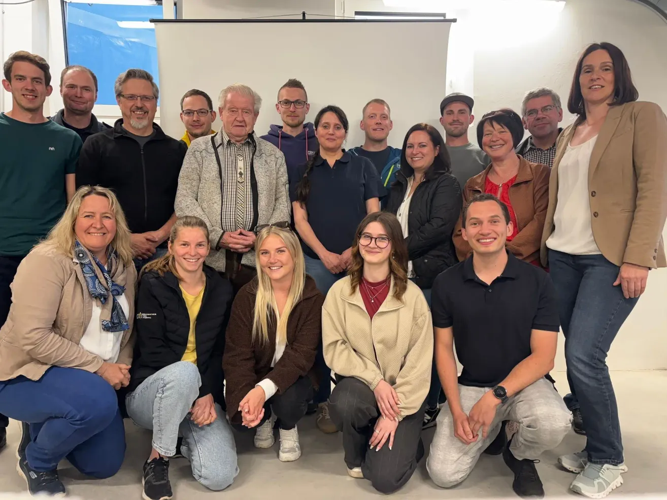 Eine Gruppe von Männern und Frauen posiert für ein Foto in einem Raum. Sie stehen und sitzen auf dem Boden, lächeln und schauen zur Kamera. Einige von ihnen tragen eine Brille. Der Raum hat eine weiße Wand, und es gibt eine blaue Abdeckung an der Wand.