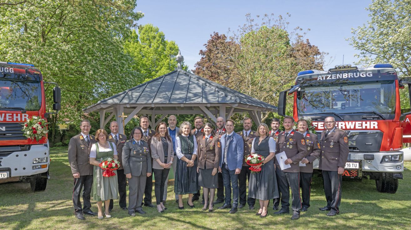 Eine Gruppe von Menschen in Uniform und ziviler Kleidung posiert für ein Foto vor einem Pavillon und einem Feuerwehrauto an einem sonnigen Tag.