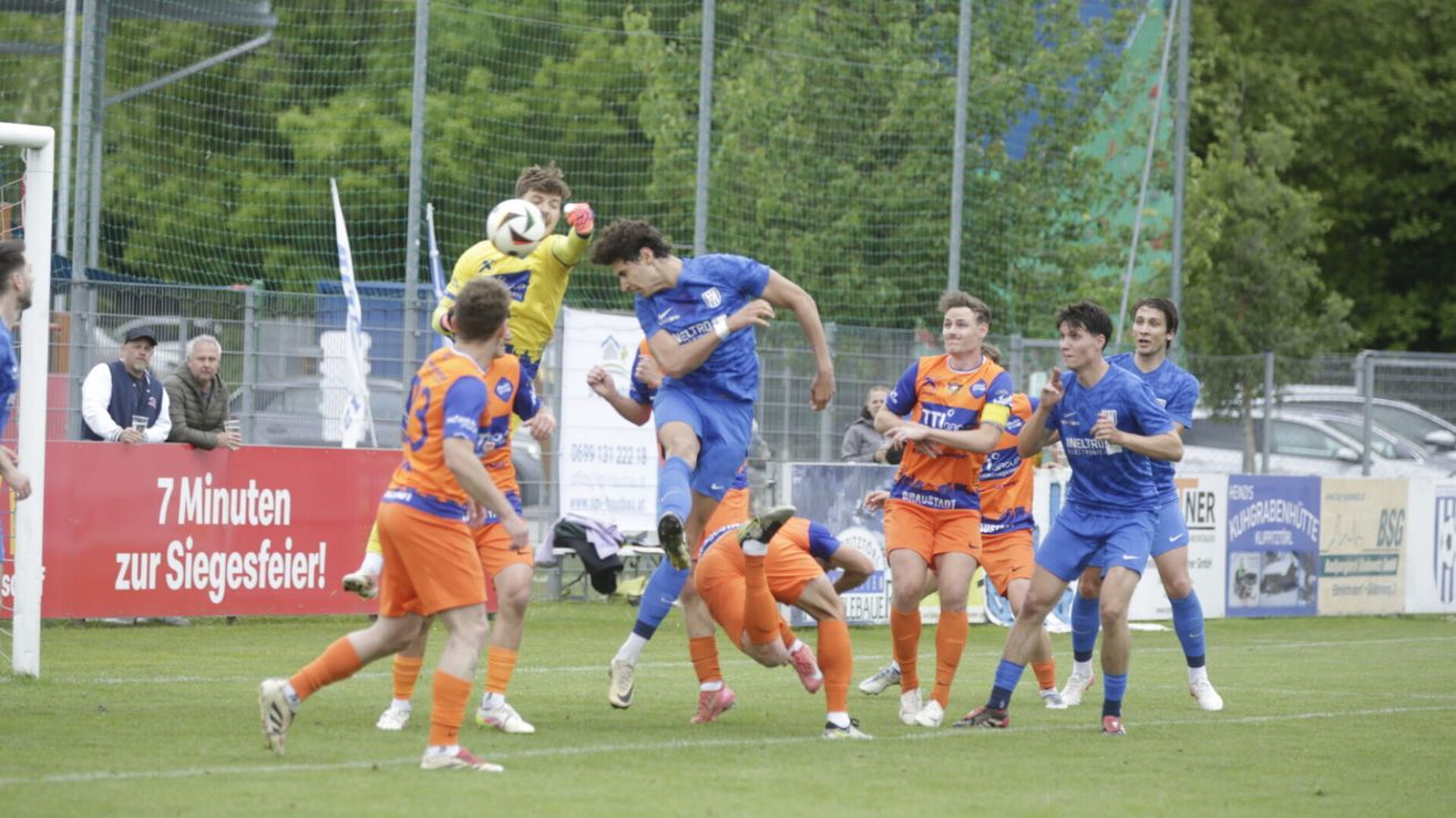 Fußballspieler in blauen und orangefarbenen Trikots sind in einem dynamischen Spiel auf einem Feld. Ein Spieler in Blau springt, um den Ball zu köpfen, während andere zuschauen.