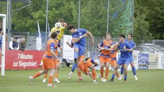 Fußballspieler in blauen und orangefarbenen Trikots sind in einem dynamischen Spiel auf einem Feld. Ein Spieler in Blau springt, um den Ball zu köpfen, während andere zuschauen.