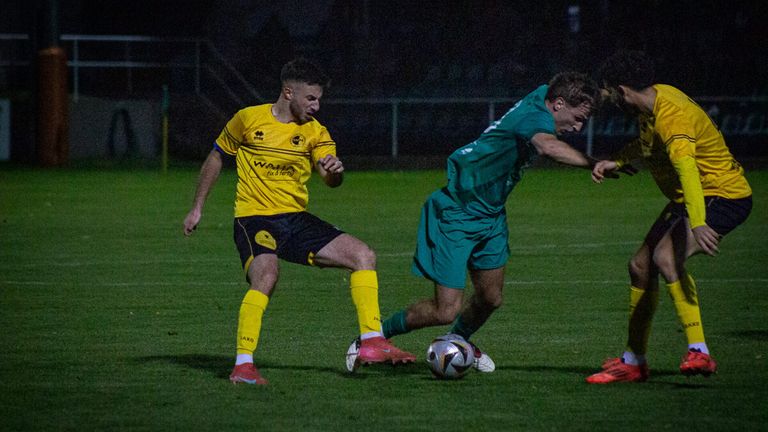 Zwei Fußballspieler spielen nachts auf einem Feld. Einer trägt ein gelbes Trikot und der andere grün. Sie jagen dem Ball hinterher.