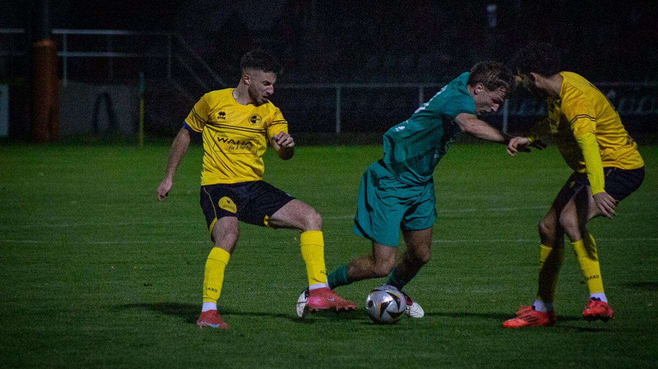 Zwei Fußballspieler spielen nachts auf einem Feld. Einer trägt ein gelbes Trikot und der andere grün. Sie jagen dem Ball hinterher.