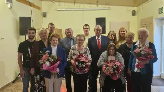 Eine Gruppe von Menschen posiert für ein Foto in einem Raum, hält Blumen und hat eine gelbe Wand, Lautsprecher und einen Projektor.