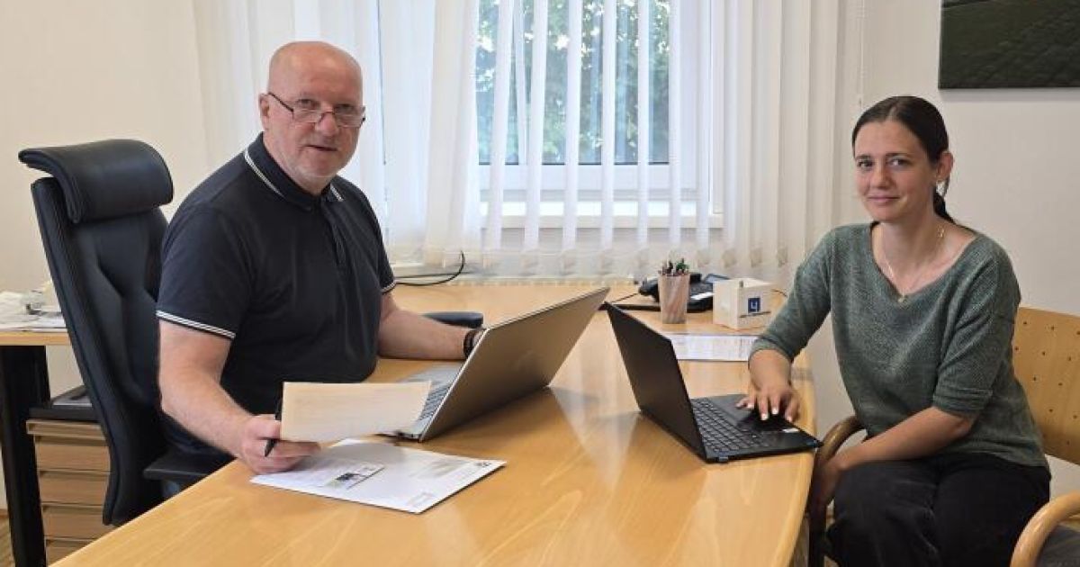 Zwei Personen sitzen an einem Schreibtisch mit Laptops und Papieren. Der Mann hält ein Papier und einen Stift, die Frau tippt auf ihrem Laptop. Im Hintergrund ist ein Fenster mit Vorhängen zu sehen.