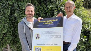 Zwei Männer halten ein Plakat mit der Aufschrift 'Wohnungsvergabe in Ansfelden.' Sie stehen vor einem grünen Busch. Das Plakat enthält einen QR-Code und einen Handlungsaufruf.