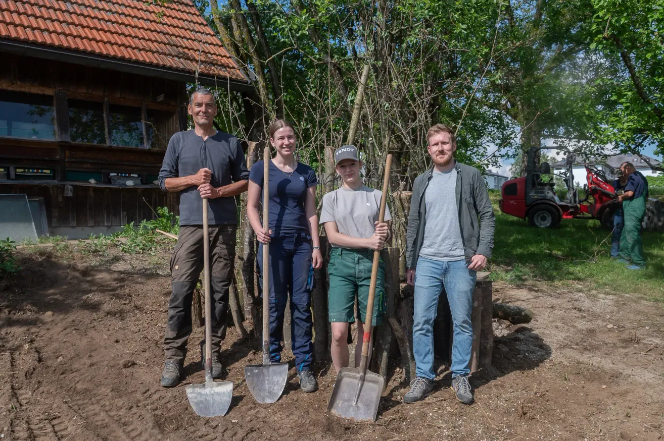 Vier Personen posieren mit Schaufeln auf einem Feld, nahe einem Haus mit rotem Dach. Sie lächeln und bereiten sich vermutlich auf Gartenarbeiten vor. Bäume und ein rotes Fahrzeug sind im Hintergrund zu sehen.