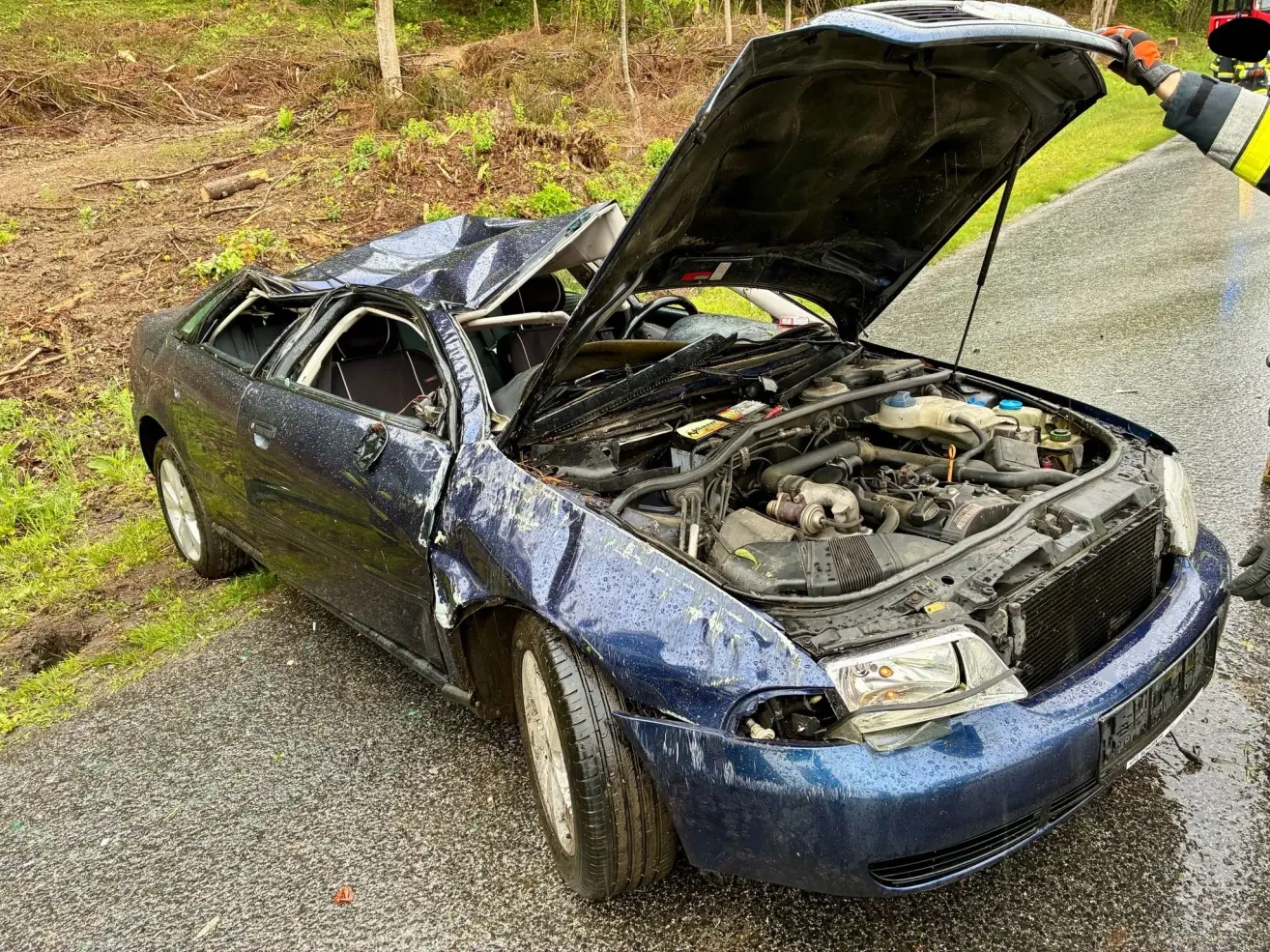 Ein blaues Auto mit einem zerbrochenen Dach steht am Straßenrand, der Motorraum ist offen. Der vordere Stoßfänger des Autos ist beschädigt.