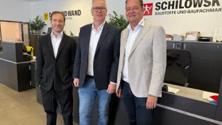 Drei Männer stehen vor einem Schreibtisch mit einer Wand dahinter, auf der 'SCHILO' steht. Der Mann links trägt einen schwarzen Anzug, der Mann in der Mitte einen Marineanzug und der Mann rechts einen hellgrauen Anzug.