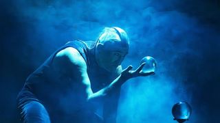 Ein älterer Mann, der eine Kristallkugel in der Hand hält, steht unter einem blauen Licht. Er schaut auf die Kugel in seiner Hand.