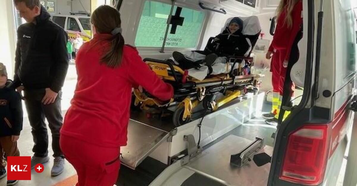 Eine Frau in Rot schiebt einen Krankenwagen-Liegenwagen mit einem Patienten, der lächelt und eine Mütze trägt. Der Liegenwagen ist auf der Rampe, und es sind zwei andere Personen in Rot.
