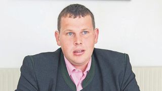 Ein Mann mit blauen Augen und einem rosa Hemd sitzt mit einer grün gefütterten Jacke an einer weißen Wand.