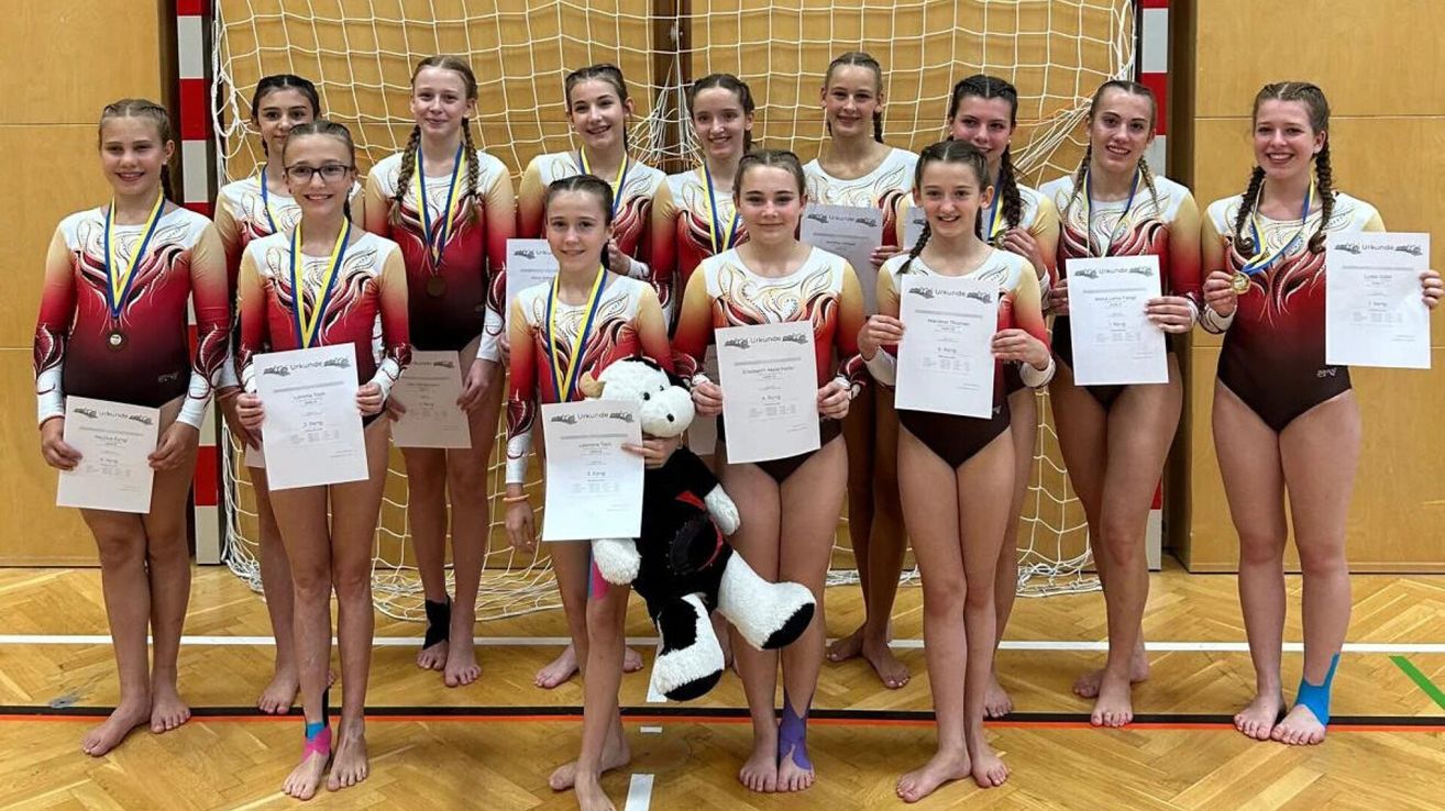 Ein Turnteam posiert für ein Foto mit Medaillen und Urkunden in der Hand. Sie stehen auf einem Holzboden vor einem Netz.