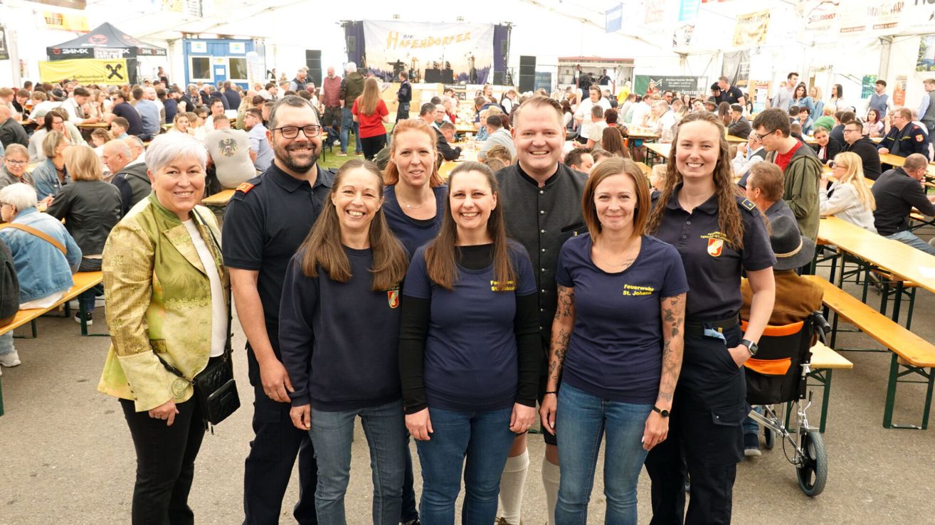 Eine Gruppe von Menschen in marineblauen T-Shirts mit Logos steht zusammen in einem Außenbereich. Dahinter sitzt eine große Menge Menschen an Tischen, wahrscheinlich bei einem Festival.