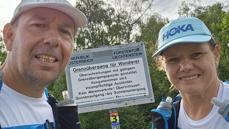 A man and a woman stand near a sign, smiling. The sign reads Grenzübergang für Wanderer, indicating a hiking trail border crossing. Trees and bushes are visible in the background.