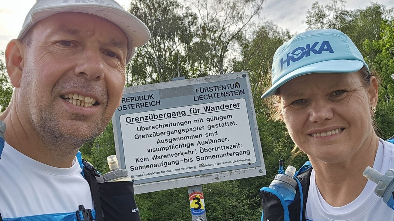 A man and a woman stand near a sign, smiling. The sign reads Grenzübergang für Wanderer, indicating a hiking trail border crossing. Trees and bushes are visible in the background.