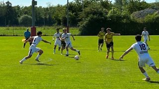 Soccer players on a field, one player is dribbling the ball while others run. The background shows trees and a clear sky.