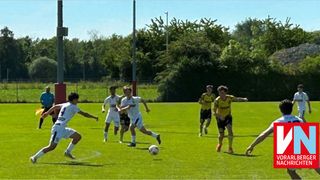 Soccer players in white and yellow uniforms are playing a match on a field with green grass and trees. One player is kicking the ball.