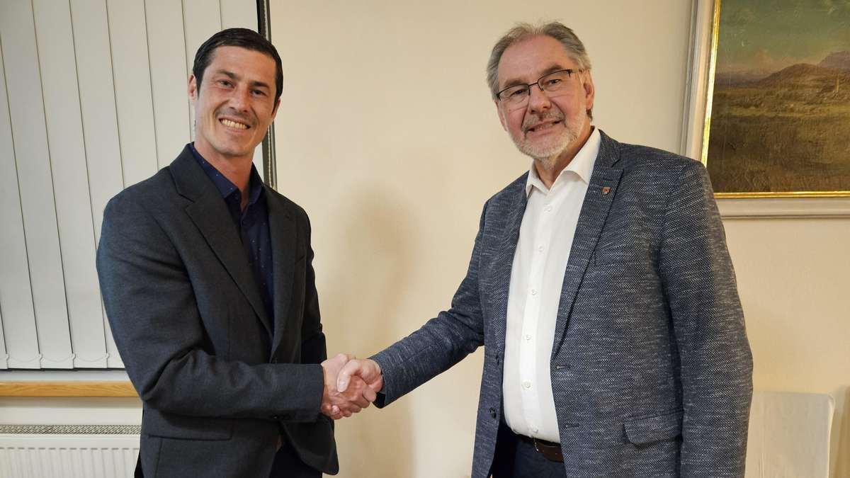 Two men in suits are shaking hands. One is younger and has a blue shirt, the other is older with gray hair and glasses, wearing a light blue coat.