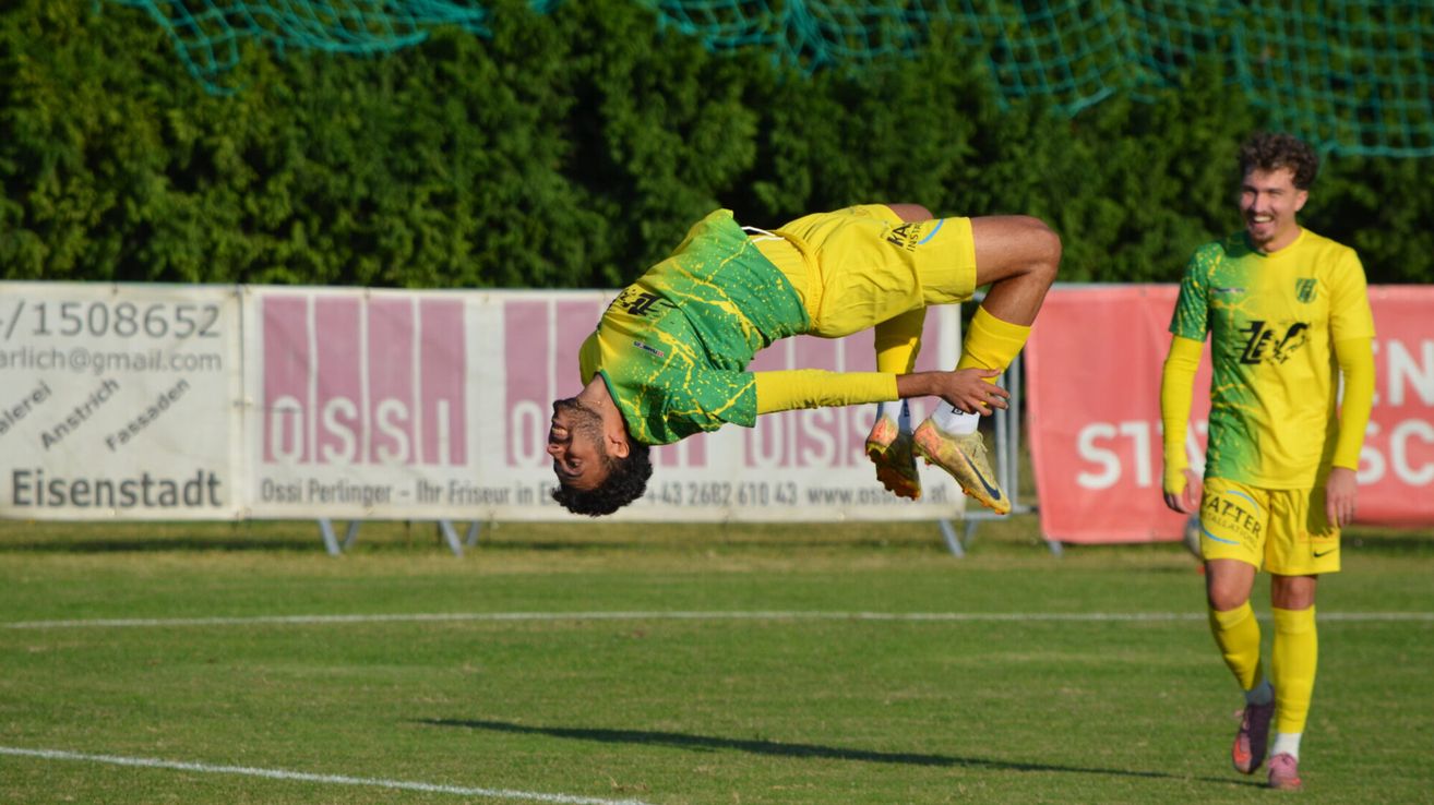 Ein Fußballspieler schwebt mit ausgestreckten Beinen in der Luft, mit einem gelben und grünen Trikot bekleidet. Im Hintergrund ist ein Fußballfeld mit einem Banner mit dem Text 'Sis' und Zahlen zu sehen.