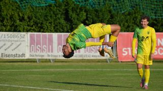 Ein Fußballspieler schwebt mit ausgestreckten Beinen in der Luft, mit einem gelben und grünen Trikot bekleidet. Im Hintergrund ist ein Fußballfeld mit einem Banner mit dem Text 'Sis' und Zahlen zu sehen.