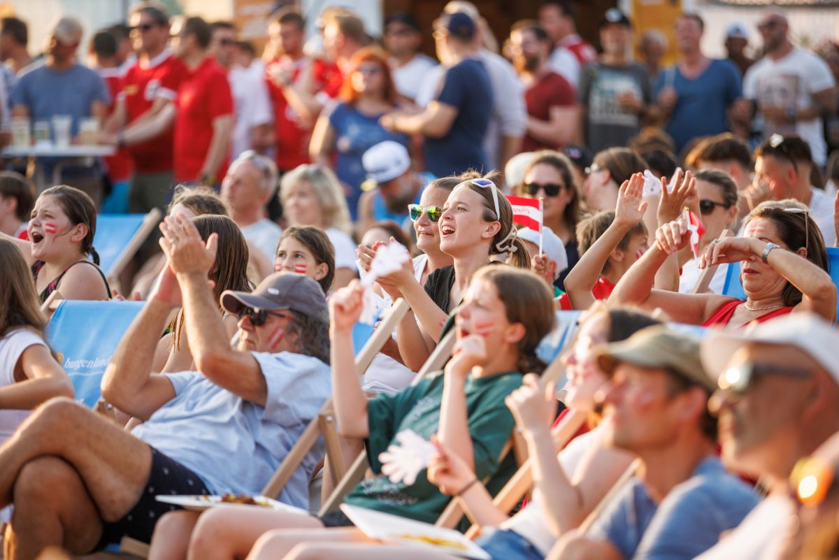 Eine Menschenmenge sitzt in einem Stadion, einige jubeln, und sie haben Flaggen und bemalte Gesichter. Das Publikum ist vielfältig, und einige tragen Hüte und Brillen. Einige halten Essen und Getränke.