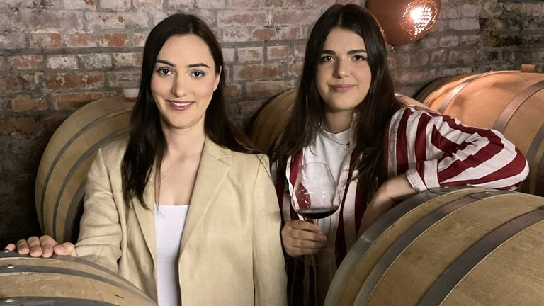 Two women are standing next to each other in a room. One woman is holding a wine glass. They are both smiling. Behind them is a brick wall and wine barrels.