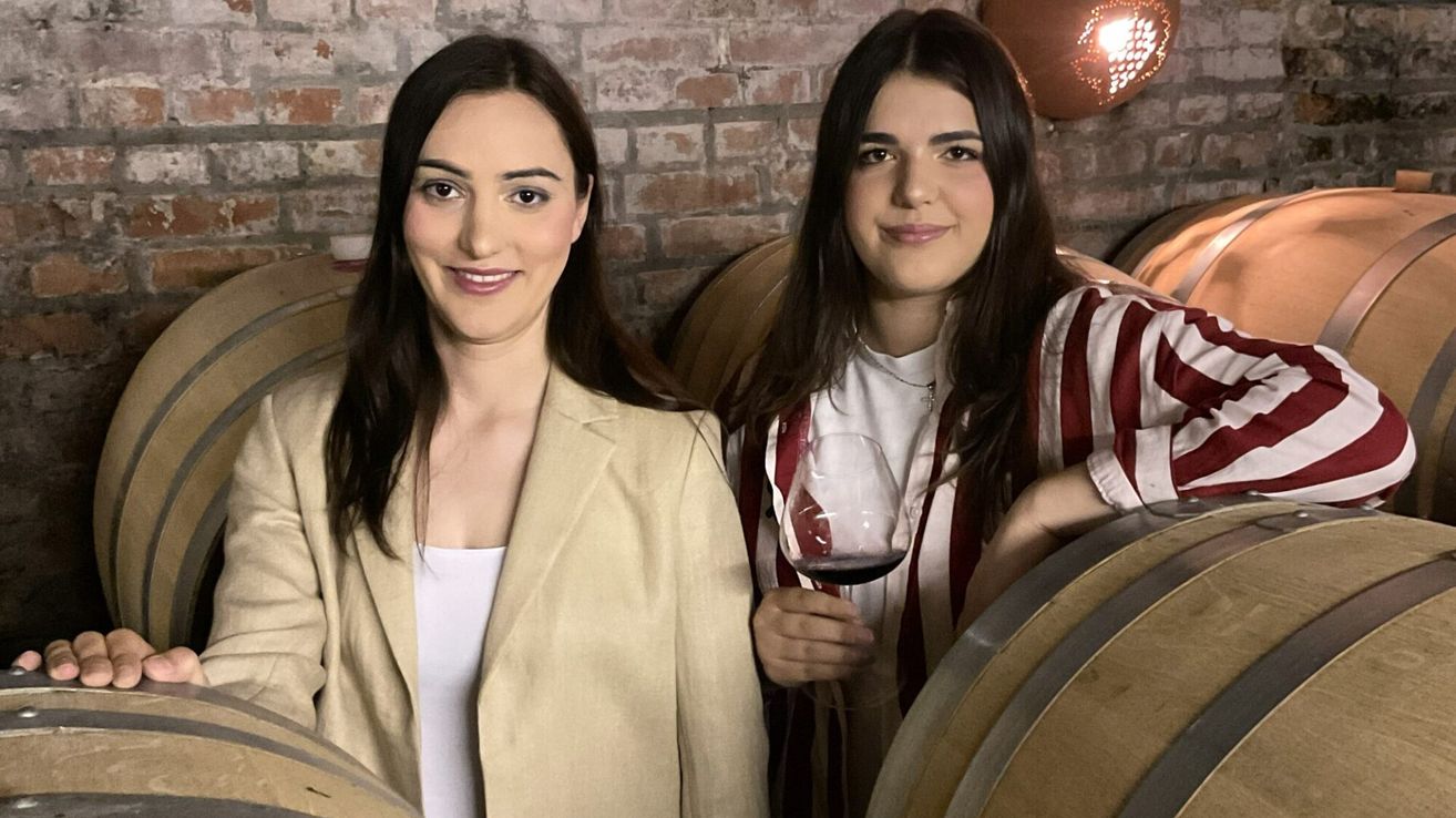 Two women are standing next to each other in a room. One woman is holding a wine glass. They are both smiling. Behind them is a brick wall and wine barrels.