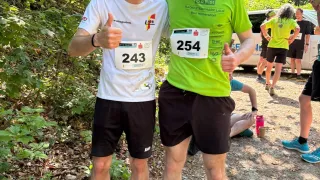 Two men stand in a forest with bibs, one showing number 243 and the other 254, giving thumbs up. Behind them are other people and a white van.