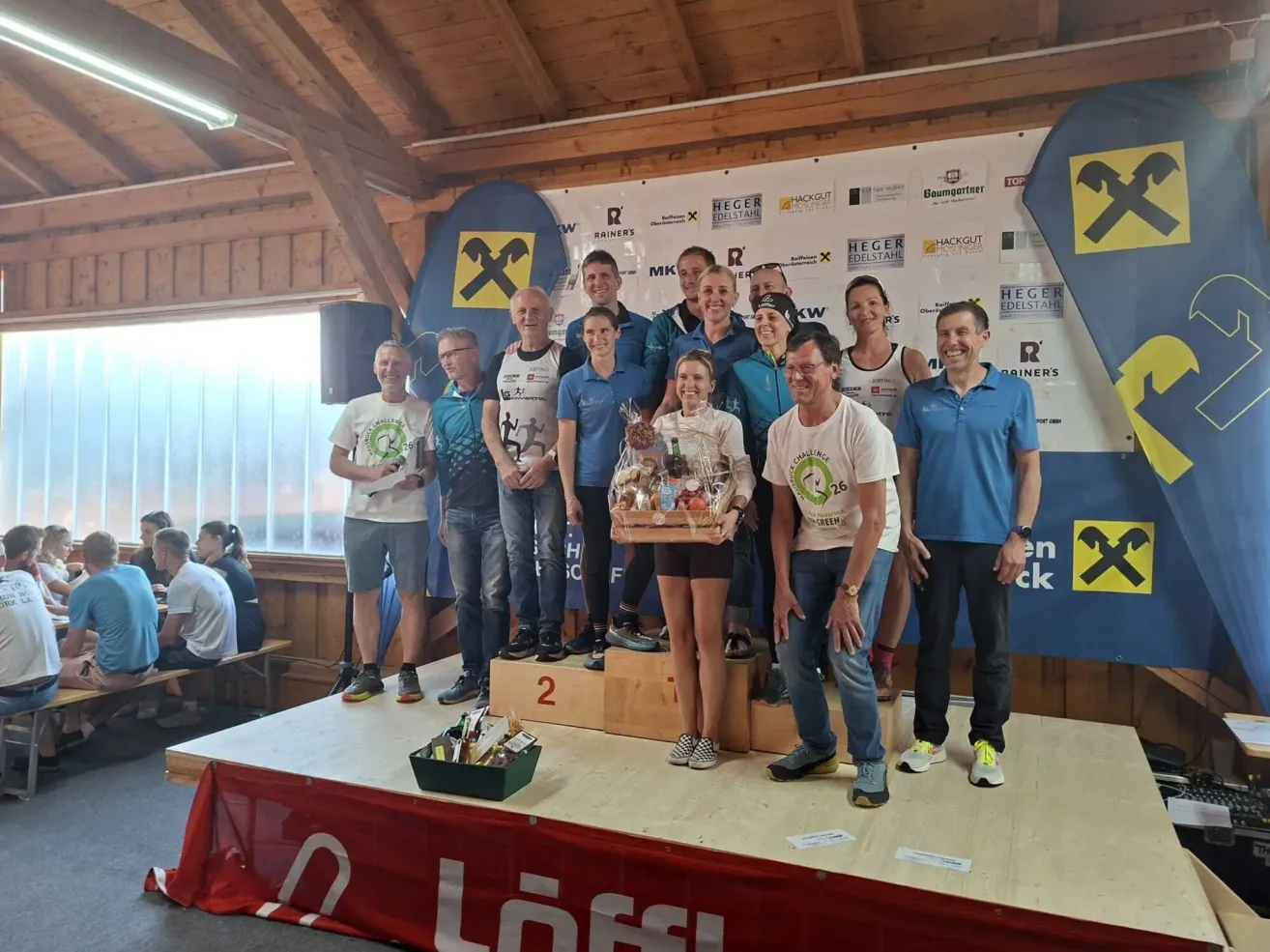 Eine Gruppe von Menschen steht auf einer Bühne, einige mit Medaillen, halten Trophäen und Geschenke, mit einem Banner dahinter und einem Banner mit dem Wort Loffl davor.