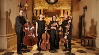 A quartet of musicians poses for a photograph in a church, each holding their respective instruments: cello, cello, violin, and sitar.