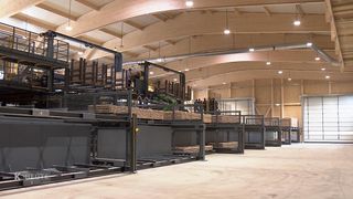 An indoor facility with multiple levels of pallet racks filled with wooden pallets. Several people are working on the racks, and some are standing in the aisle. The ceiling is high and made of wood.